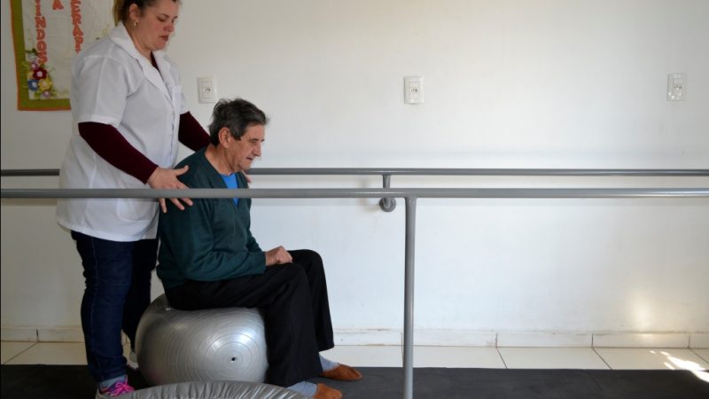 Fisioterapeuta utiliza bola para trabalhar equilíbrio e coordenação com seu Antoninho.  